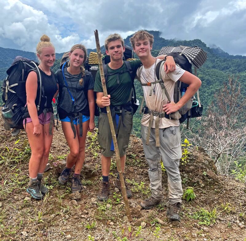 Four young hikers pose for a photo on a mountain trail. They are equipped with large backpacks and hiking gear, suggesting they are on a multi-day trek. The background features a lush, green mountainous landscape under a cloudy sky. The hikers appear to be taking a break, standing close together and smiling at the camera.